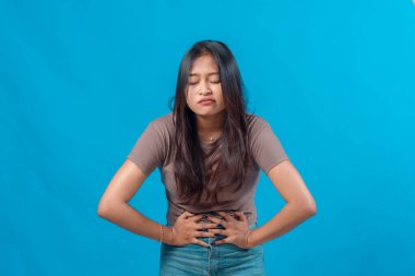 Karın ağrısı ya da rahatsızlığı olan genç bir Asyalı kadın, iki eli ve gözü kapalı karnını tutuyor, acı verici ya da hastalıklı bir duygu ifade ediyor, mavi arka planda izole edilmiş..