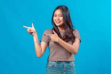 Genç Asyalı kadın, iki elini de kenarda göstererek güvenle gülümsüyor, metin ya da ürün yerleştirme için yer gösteriyor, mavi arkaplan önünde duruyor..