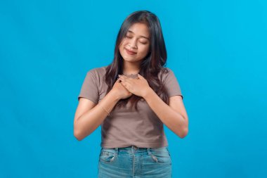 Genç Asyalı kadın gözlerini kapatıp göğsüne sarıldı. Minnettarlığını ya da minnettarlığını ifade etti. Mavi arka planda sakince durdu..