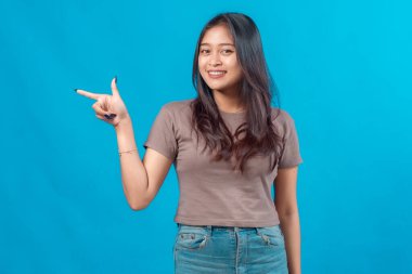 Sıradan kıyafetlerle gülümseyen genç bir Asyalı kadının portresi işaret parmağıyla yan tarafı gösteriyor, mavi arka planda güvenle duruyor, reklam konsepti için mükemmel..