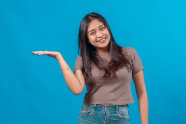 Neşeli genç Asyalı kadın gülümsüyor ve tek eliyle boşluğu tutuyor, ürün konseptini sunuyor ya da gösteriyor. Mavi arka planda stüdyo çekimi, reklamlar için mükemmel..