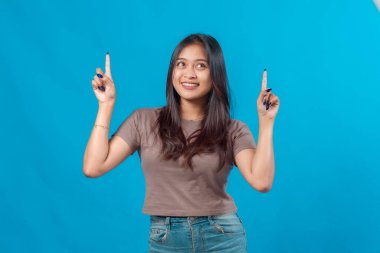 Genç Asyalı kadın gülümsüyor ve işaret parmaklarını yukarı kaldırarak parlak mavi bir arkaplana bakıyor. Reklam, pazarlama ve promosyon için mükemmel..