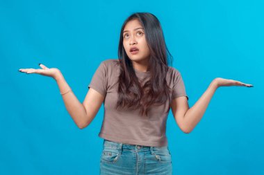 Kafası karışmış genç Asyalı kadın tişört ve kot pantolon giymiş, elleri havada, yukarı bakıyor belirsizlikle, mavi arka planda izole edilmiş, kafa karışıklığı ya da şüphe kavramı.