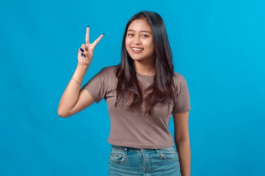 Gündelik kıyafetlerle gülümseyen Asyalı genç bir kadın barış ya da zafer el hareketi sergiliyor, parlak mavi arka plana karşı güvenle duruyor. Arkadaş canlısı, neşeli ve anlamlı bir portre konsepti..