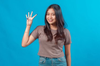 Normal kıyafetli gülümseyen genç Asyalı kadın, dört parmağını sağ eliyle gösteriyor, düz mavi bir arka planın önünde duruyor, neşeli bir ifadeyle kameraya bakıyor..