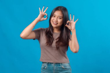 Gülümseyen Asyalı genç bir kadın iki eliyle iki kere el hareketi yapıyor, pozitifliğini ve onayını ifade ediyor, mavi arka planda kendine güvenen bir şekilde duruyor..
