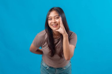 Genç Asyalı kadın, parlak bir şekilde gülümsüyor ve elleri ağzının kenarında bağırıyor, parlak mavi arka plana karşı günlük kıyafetler içinde öne eğiliyor. Stüdyo çekimi, neşeli ifade.