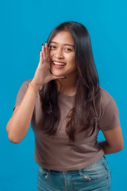 Neşeli genç Asyalı kadın, eli ağzının yanında gülümseyip bağırıyor, parlak mavi arka planda günlük kıyafetler giyiyor. Stüdyo görüntüsü, kopyalama alanı.