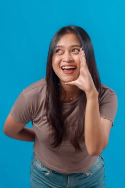 Neşeli genç Asyalı kadın, eli ağzının kenarında gülümseyip bağırıyor, yukarı bakıyor, heyecan ve neşe ifade ederken stüdyoda parlak mavi arka planda duruyor..