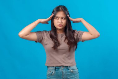 Genç Asyalı kadın şaşkın ve bunalmış hissediyor, iki elini yana bakarken kaldırıyor, sağlam mavi bir arka plana karşı duruyor ciddi bir ifadeyle..