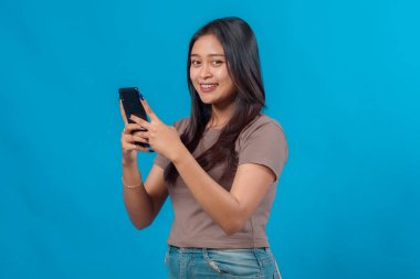 Kameraya bakarken akıllı telefon kullanan gülümseyen genç Asyalı kadın, düz mavi bir arka plan karşısında duruyor, günlük tişört ve kot pantolon giyiyor, neşeli ve modern..
