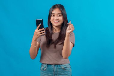 Akıllı telefonu tutarken gülümseyen ve Koreli genç Asyalı bir kadın. Parmaklarıyla kalp hareketi yapıyor. Mavi arka planlı stüdyo portresi, aşkı, neşeyi ve bağlantıyı simgeliyor..