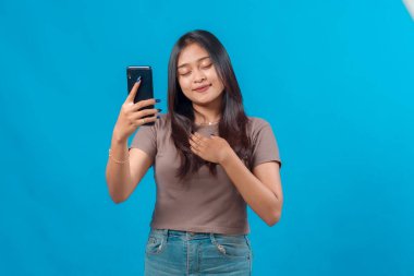 Genç Asyalı bir kadın barışçıl bir gülümseme ve göğsü olan bir selfie çekiyor, gözleri minnettarlıkla kapalı, stüdyo ışıklarında parlak mavi bir arka plana karşı duruyor..