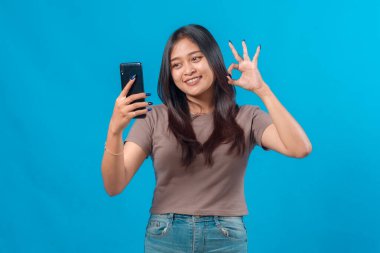 Neşeli genç Asyalı kadın akıllı telefonuyla selfie çekerken normal bir el hareketi gösteriyor. Mavi arka planda dikiliyor, günlük kıyafetlerini giyiyor..