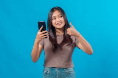 Akıllı telefon tutan ve başparmağını kaldıran kendine güvenen genç bir Asyalı kadın, başparmağıyla başka tarafa bakıyor, günlük kıyafetler giyiyor, mavi bir arka plana yaslanıyor, parlak bir şekilde gülümsüyor..