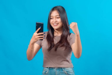 Genç Asyalı kadın akıllı telefonuyla selfie çekerken gülümsüyor ve başarılı bir yumruk hareketi yapıyor. Mavi arka plan karşısında başarıyı kutluyor..