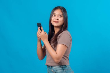 Genç bir Asyalı kadın, elinde akıllı bir telefonla gülümsüyor, düz mavi bir arkaplan karşısında duruyor. Düşünceli bir ifadeyle yana bakıyor..