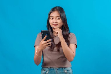 Kendine güveni tam genç Asyalı bir kadın akıllı bir telefon tutarken çenesine özenle dokunuyor. Stüdyoda mavi bir arka planda duruyor..