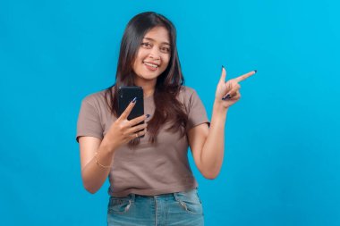 Neşeli, genç Asyalı bir kadın elinde akıllı telefon tutarken gülümseyip yan tarafı işaret ediyor, bir şeyleri teşvik ediyor ya da tavsiye ediyor, parlak mavi arka planda izole edilmiş..