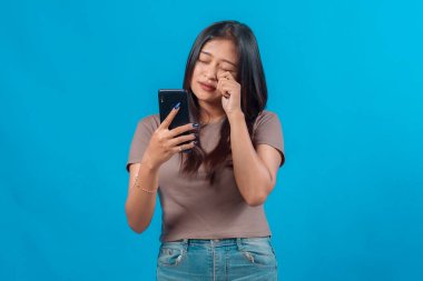 Genç Asyalı bir kadın akıllı telefonuna bakıyor. Gözleri kapalı ve nazikçe bir gözünü ovalıyor. Yorgunluğunu ya da duygusal tepkisini ifade ediyor..