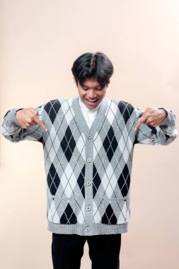 Asyalı genç adam iki parmağıyla işaret ederken gülümsüyor, baklava desenli bir süveter giyiyor, neşeli bir ifade, güven ve reklamcılık ve yaşam tarzı kavramları için pozitif bir tutum sergiliyor..