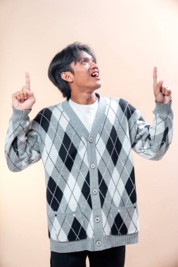 Gülümseyen Asyalı genç bir adam, desenli bir süveter giyiyor, iki parmağıyla yukarıya bakıyor, neşeli bir ifade sergiliyor, iş için mükemmel, yaşam tarzı, ve reklam temaları..