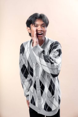 Genç Asyalı bir adam gri bir baklava desenli süveter giyiyor, bir eli ağzının yanında poz veriyor bağırıyor ya da çağırıyor gibi, iletişim kuruyor, anons ediyor ve modern bir yaşam tarzında dikkat çekiyor..
