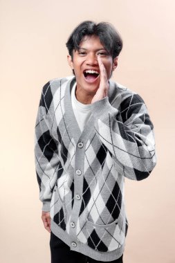 Asyalı adam gri baklava desenli hırka giymiş, neşeli bir yüz ifadesiyle elini ağzının yanında tutarak bağırıyor, canlı ve kendinden emin bir stil sergiliyor..