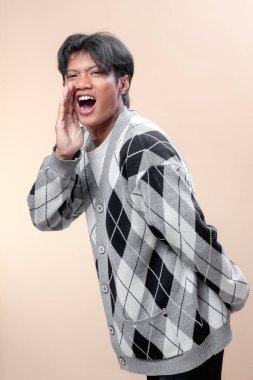 Asyalı bir adam, gri baklava desenli hırka giyiyor, eli ağızda bağırıyor, enerjik bir ifade sergiliyor, kendine güvenen bir tavır takınıyor ve güçlü bir iletişim hareketi sergiliyor..