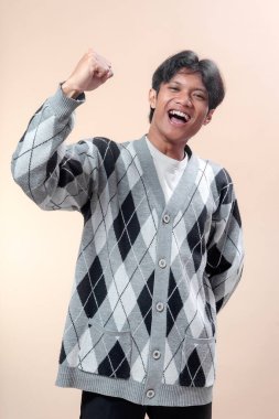 Neşeli, genç Asyalı bir adam parlak bir şekilde gülümsüyor, gri bir baklava desenli süveter giyiyor, heyecanla yumruğunu kaldırıyor, kendine güvenini, neşesini, iyimserliğini ve başarısını gösteriyor..