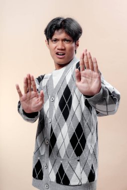 İfade veren yüz ifadesi olan Asyalı adam, çift el hareketi gösteriyor, gri bir baklava desenli süveter giyiyor, reddedilmeyi sembolize ediyor, uyarı, savunma, anlaşmazlık ve iletişimde ihtiyatı..
