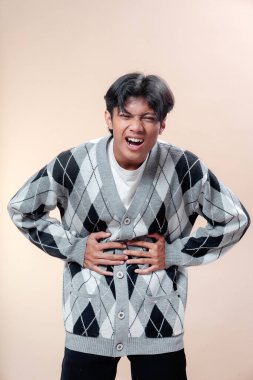 Asyalı bir adam gri baklava desenli süveter giyiyor. İki eliyle karnını tutarken acı içinde kıvranıyor. Dramatik bir duruşta güçlü bir duygu ve rahatsızlık gösteriyor..