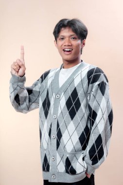Asyalı bir adam gri baklava desenli süveter giyer. Tek parmağıyla yukarı doğru gülümserken pozitif bir ifade, fikir konsepti ve neşeli bir tavır sergiler..