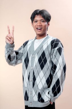 Gri baklava desenli süveter giyen Asyalı adam gülümserken zafer el hareketini iki parmağıyla gösteriyor, mutluluğu, güveni ve pozitif neşeli tavırları ifade ediyor..