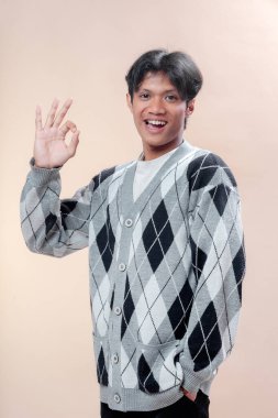Gri baklava desenli süveter giyen Asyalı genç bir adam el hareketini, onayı, güveni ve olumlu tutumu sembolize ederken parlak bir şekilde gülümsüyor..