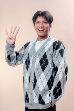 Asyalı adam gri baklava desenli süveter içinde gülümserken tek eliyle dört parmağını kaldırıyor, neşeli tavır, kendine güven, olumlu duygu ve genç profesyonel görünüm sergiliyor..