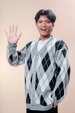 Tek eliyle beş parmağını kaldırırken gülümseyen gri baklava desenli süveter giyen Asyalı adam neşeli bir jest, pozitif tavır, kendine güven, gençlik ruhu ve dost canlısı profesyonel imaj sergiliyor..