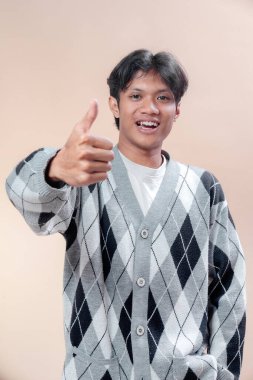 Gri baklava desenli süveter giyen genç bir Asyalı, onay, güven, motivasyon ve başarıyı sembolize eden bir jest yaparken parlak bir şekilde gülümsüyor..