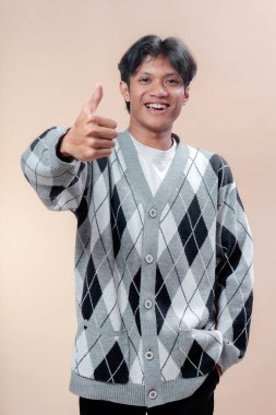 Gri baklava desenli süveter giyen Asyalı genç bir adam başparmağıyla onaylama, pozitiflik ve profesyonel tutumu sembolize ederken kendinden emin bir şekilde gülümsüyor..
