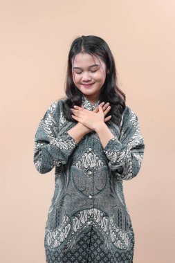 Geleneksel batik giyinen Asyalı kadın ellerini göğsüne koyuyor. Huzur içinde gülümsüyor. Minnettarlık, güven, kültürel gurur ve profesyonel tavır sergiliyor..