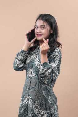 Zarif batik elbiseler giyen Asyalı kadın akıllı telefonla konuşurken gülümsüyor, parmak hareketi gösteriyor, sevgiyi, kültürü ve bağlantıyı simgeliyor..