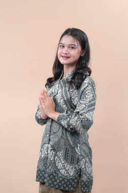 Batik elbise giymiş genç bir Asyalı kadın saygılı bir selamlama jesti ile poz verirken sıcak bir şekilde gülümsüyor, zarafet, kültür ve profesyonellik gösteriyor..
