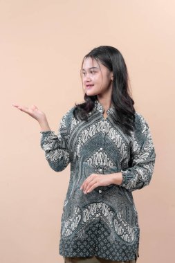 Zarif batik kıyafetler giyen Asyalı bir kadın, kendine güvenen bir gülümsemeyle poz veriyor, eliyle işaret ediyor, cazibesini ve profesyonel ifadesini gösteriyor..