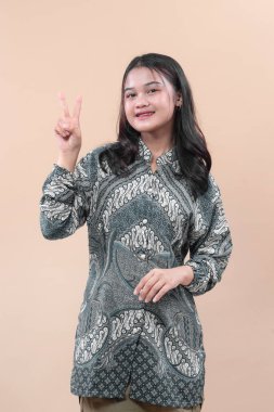 Zarif batik giysiler içinde Asyalı bir kadın parmaklarıyla barış işareti gösterirken gülümsüyor, kendine güvenini, çekiciliğini, pozitifliğini ve profesyonel tarzını ifade ediyor..