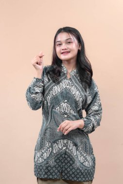 Zarif batik giysiler giyen genç Asyalı kadın parmak kalp hareketi yapıyor, içten gülümsüyor, küresel reklam ihtiyaçları için neşe, pozitiflik, kültür ve profesyonelliği ifade ediyor..
