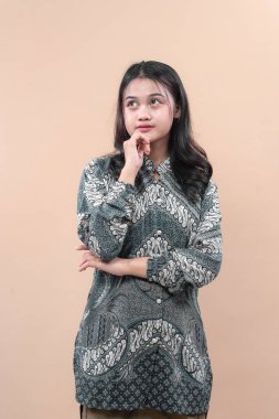 Asyalı bir kadın geleneksel batik gömlek giyiyor. Düşünceli bir ifadeyle poz veriyor. El çeneye dokunuyor ve yukarı bakıyor..