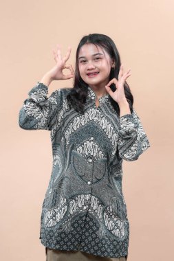 Geleneksel batik gömlek giyen Asyalı bir kadın, iki el hareketi yaparken kendine güvenerek gülümsüyor, sade arka planda neşeli bir ruh hali sergiliyor..