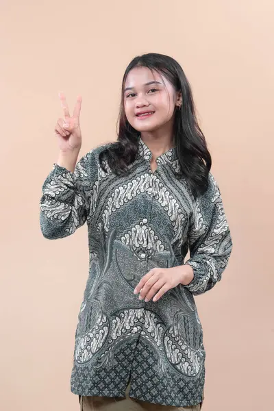 Zarif batik giysiler içinde Asyalı bir kadın parmaklarıyla barış işareti gösterirken gülümsüyor, kendine güvenini, çekiciliğini, pozitifliğini ve profesyonel tarzını ifade ediyor..