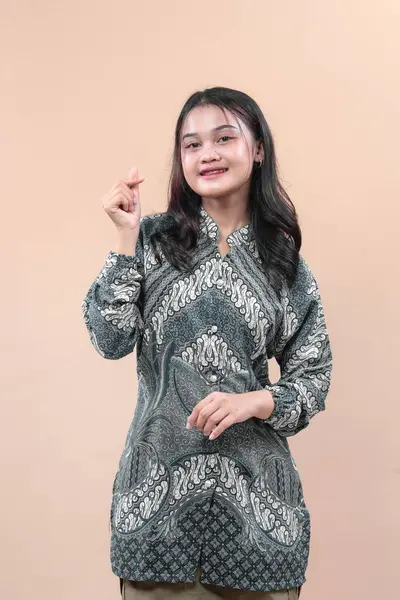 Zarif batik giysiler giyen genç Asyalı kadın parmak kalp hareketi yapıyor, içten gülümsüyor, küresel reklam ihtiyaçları için neşe, pozitiflik, kültür ve profesyonelliği ifade ediyor..