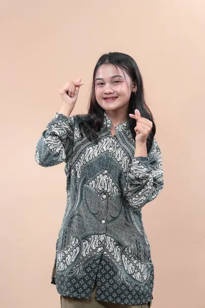 Zarif batik tişört giyen gülümseyen genç Asyalı kadın iki eliyle parmak kalp hareketi yapıyor. Küresel reklam kavramları için mutluluk, güven ve kültürel gurur gösteriyor..